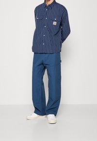 Mies, joka on pukeutunut tummansinisiin pystysuoriin raidallisiin nappipaitaan, löysiin sinisiin housuihin ja valkoisiin lenkkareihin, seisoo valkoista taustaa vasten.