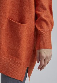 Oranžový pletený svetr s měkkou texturou, vybavený přední kapsou, žebrovanými manžetami a bočními rozparky, fotografovaný na neutrálním pozadí.