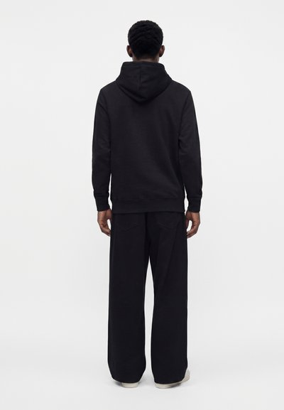 Personne debout, dos à la caméra, portant un sweat à capuche noir et un pantalon noir large sur un fond clair uni.