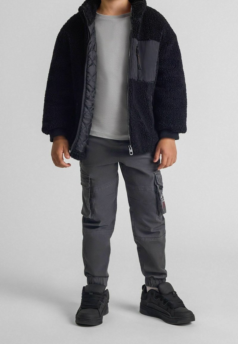Enfant portant une veste polaire noire par-dessus un t-shirt gris à manches longues, un pantalon cargo foncé et des baskets noires sur un fond uni.
