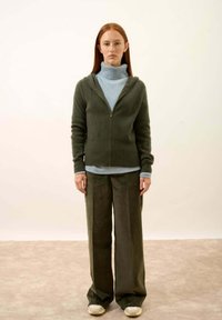 Sweater zippé vert olive avec une finition texturée, porté par-dessus un col roulé bleu clair. Pantalon baggy vert foncé et chaussures beiges.