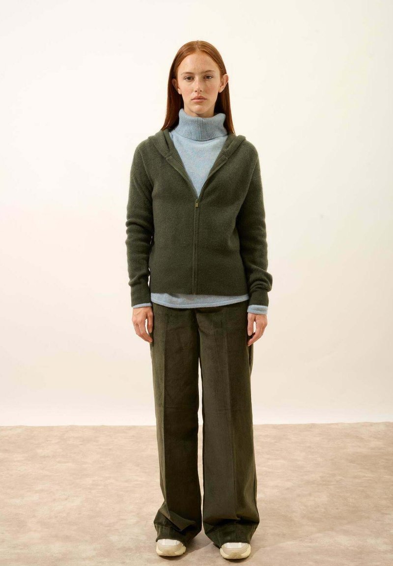 Sweater zippé vert olive avec une finition texturée, porté par-dessus un col roulé bleu clair. Pantalon baggy vert foncé et chaussures beiges.