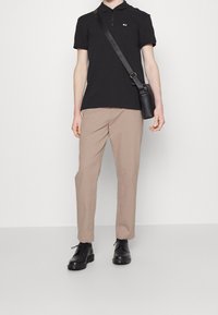 Sort poloshirt med lynlåsåbning i kraven, beige bukser og sorte pælsko. Modellen holder en lille sort skuldertaske.