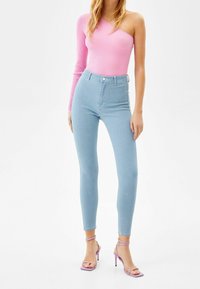 Jeans de tiro alto en azul claro combinados con una camiseta de canalé rosa de un solo hombro y sandalias moradas de tacón alto con tiras. Texturas suaves, diseño ajustado.