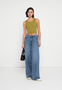 Κροπ τοπ σε ελιά πράσινη απόχρωση συνδυασμένο με τζιν παντελόνι ψηλόμεσο, με φαρδιά πόδια σε μπλε χρώμα. Η μοντέλο κρατάει μια μαύρη τσάντα χειρός. Απλός, casual σχεδιασμός.