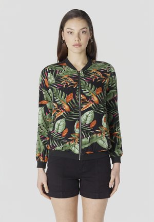 Mujer con chaqueta negra de cremallera con patrones de hojas tropicales verdes y naranjas y pantalones cortos negros, de pie contra un fondo claro y liso.