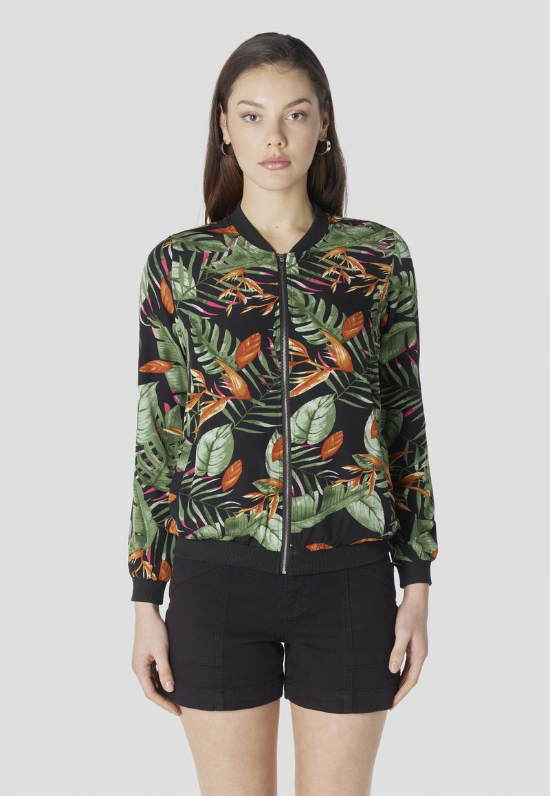 Vrouw met een zwarte jas met ritssluiting met groene en oranje tropische bladeren en zwarte shorts, staand tegen een effen lichte achtergrond.