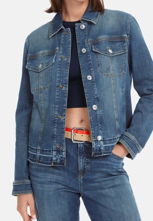 Vrouw die een blauwe spijkerjas en jeans draagt met een zwart crop top, hand in de zak en een rood-beige riem zichtbaar rond de taille.