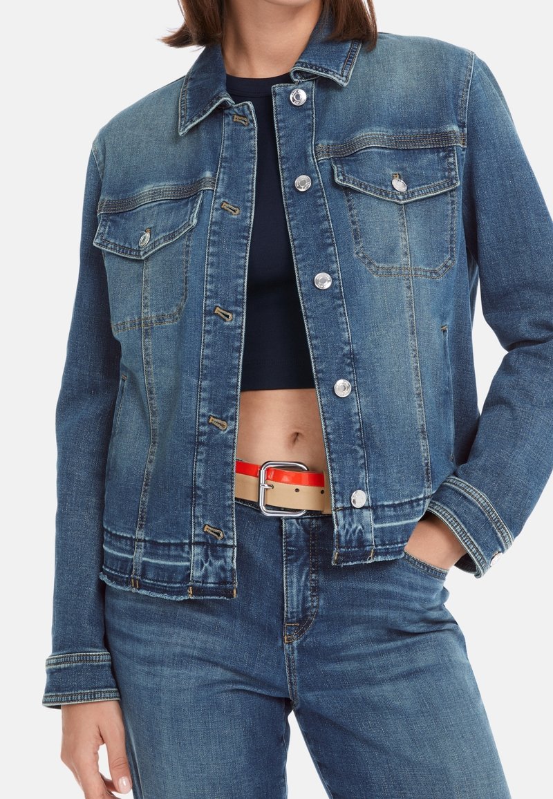 Femme portant une veste en jean bleue et un jean avec un crop top noir, main dans la poche, et une ceinture rouge et beige visible autour de la taille.