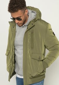 Giacca puffer verde oliva con finitura testurizzata, dotata di cappuccio con coulisse, tasche con zip e dettagli in bottone sopra un hoodie grigio.