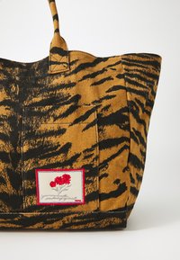 Canvas-Tasche mit einem mutigen, schwarzen Tigerstreifenmuster auf einem senfgelben Hintergrund. Mit einem rechteckigen Aufnäher mit roten Blumen und weißen Details.