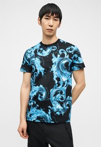 Άνδρας που φοράει μαύρο μπλουζάκι με έντονα μπλε κυματιστά σχέδια, στέκεται μπροστά από ένα απλό φωτεινό φόντο.