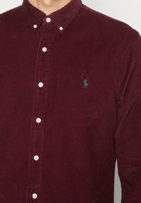 Burgundská kordurová košile s límečkem na knoflíky, bílými knoflíky a malým zeleným logem polo na hrudi. Textura obsahuje žebrovaný vzor.