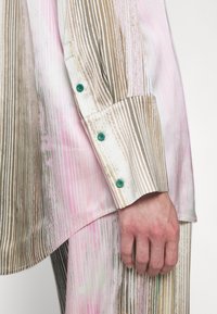 Chemise à boutons avec des rayures verticales en rose, marron et blanc, d'une texture lisse et avec des boutons verts sur les poignet.