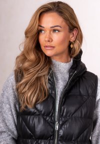 Woman with long wavy blonde hair wearing gold hoop earrings, a grey sweater, and a zipped black quilted vest, looking to her left.
