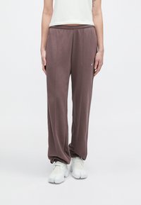 Pessoa vestindo calças de moletom cor malva com punhos franzidos e tênis brancos, em pé contra um fundo branco liso.