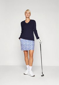 Tengerészkék bordázott kötött pulóver V-nyakkal, mintás kék-fehér szoknyával, fehér tornacipővel és golfütőt tartva.