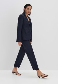 Costume en lin bleu marine avec un blazer ajusté et un pantalon droit. Porté avec des sandales noires à talons, au design minimaliste et aux lanières.