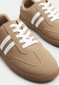 Una coppia di sneakers beige con lacci bianchi e due strisce bianche sui lati, dotate di suole marroni texture.