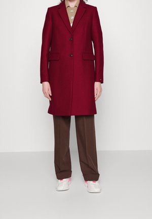Person iført en lang vinrød frakke over en beige knappet skjorte, brune bukser og hvide sneakers med pink detaljer, stående mod en ensfarvet baggrund.