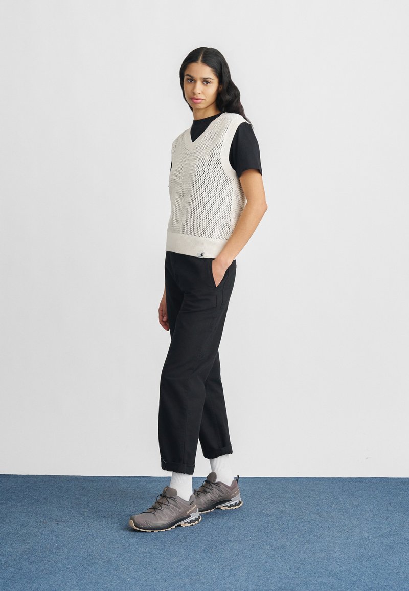 Top smanicato in maglia color crema con scollo a V, abbinato a pantaloni neri e scarpe sportive grigie. Il modello posa su un tappeto blu davanti a uno sfondo neutro.