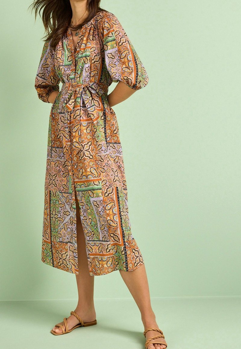 Femme debout, les mains derrière le dos, portant une robe midi à motifs avec des manches bouffantes et des sandales beiges sur un fond vert clair.
