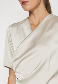 Femme portant un haut cache-cœur en satin beige clair à manches courtes, montré de l'épaule au menton.