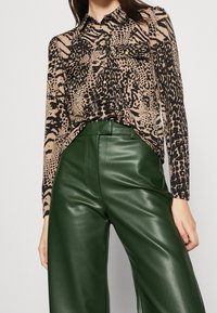 Dierafdruk blouse in beige en zwart, gecombineerd met hoog-geprijsde groene neplederen wijde broek. Blouse heeft een knoopsluiting aan de voorkant.