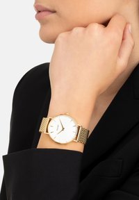 Frau mit einer goldfarbenen Mesh-Armbanduhr mit weißem Zifferblatt ohne Zahlen, gekleidet in einem schwarzen Blazer.