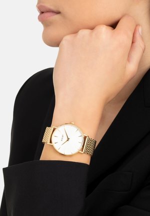 Frau mit einer goldfarbenen Mesh-Armbanduhr mit weißem Zifferblatt ohne Zahlen, gekleidet in einem schwarzen Blazer.