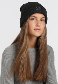 Black knitted beanie hat featuring a small white Puma logo tag. The model wears a gray ribbed sweater, with long hair flowing down.