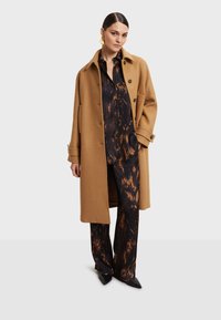 Smeđa vunena kaput s velikim ovratnikom, zatvaranje na gumbe. Tamnoslikana bluzica i široke hlače, uparene s crnim cipelama s špicastim nosom.