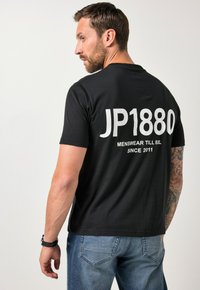 Czarny t-shirt z krótkim rękawem z dużym białym napisem "JP1880" na plecach oraz frazą "MENSWEAR TILL 8XL SINCE 2011."