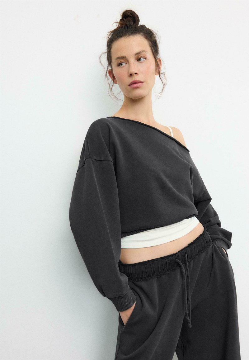 Jeune femme avec un chignon, portant un sweat-shirt noir ample à épaules dénudées, un débardeur blanc et un pantalon de jogging noir, les mains dans les poches, regardant sur le côté.