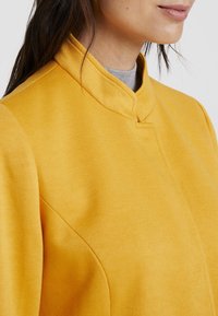 Sininen sinappi keltainen takki, jossa on teksturoitu pinta, mandariinikaulus ja hienovaraiset sauma-yksityiskohdat, esittelee minimalistista muotoilua.