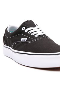 Musta kangaskengät valkoisilla nauhoilla, valkoisella kumipohjalla ja mustalla ommella. Sivussa on pieni "VANS"-merkki.