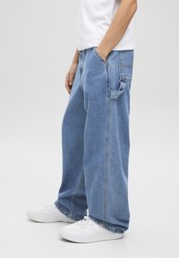 Jeans larghi di denim di un blu chiaro, con un classico design a cinque tasche e dettagli di cucitura visibili. Indossati con sneakers bianche.