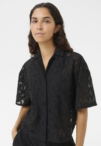 Chemise noire à manches courtes avec un motif texturé en tissage ouvert, deux poches avant, un col à encoche et une coupe ample.