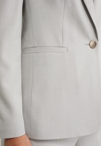 Světle šedý blazer s hladkou texturou, zapínáním na jeden knoflík a boční kapsou. Látka vypadá jemně tkaná a dobře ušitá.