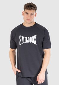 T-paita lyhyillä hihoilla, tummanharmaata pehmeää kangasta, jossa on rohkeaa valkoista tekstiä "SMILODOX" edessä. Yksinkertainen, rento istuvuus.