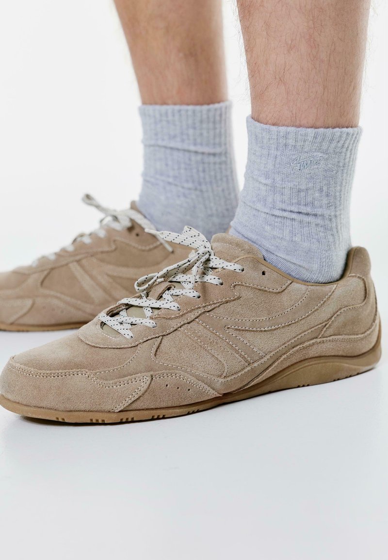 Beżowe zamszowe sneakersy z białymi teksturowanymi sznurówkami, o niskim fasonie i perforowanymi detalami po bokach, w zestawieniu z jasnoszarymi skarpetami z prążkami.
