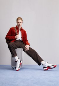 Veste en velours côtelé rouge, t-shirt blanc, pantalon marron et baskets blanches avec des accents rouges. La personne est assise sur une valise métallique.