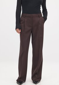 Braune, weit geschnittene Hose mit Falten, die eine glatte Textur und eine maßgeschneiderte Passform aufweist. Kombiniert mit einem enganliegenden schwarzen Langarmoberteil und eleganten schwarzen Schuhen.