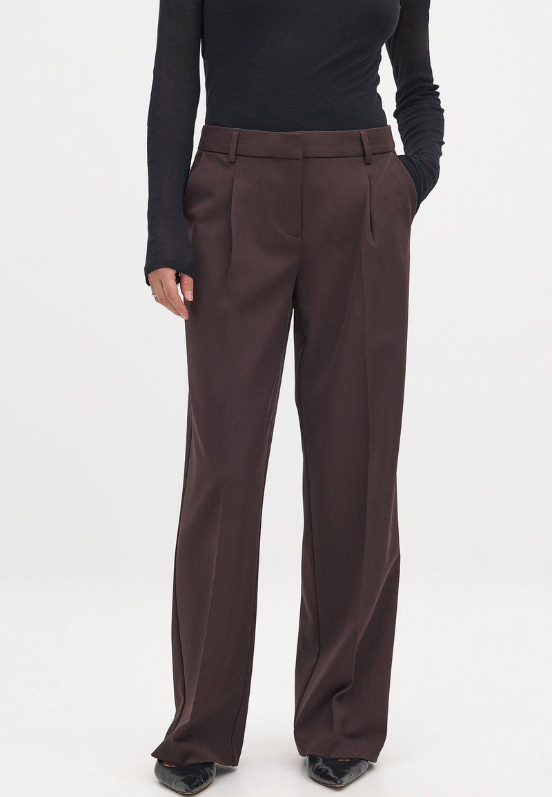 Braune, weit geschnittene Hose mit Falten, die eine glatte Textur und eine maßgeschneiderte Passform aufweist. Kombiniert mit einem enganliegenden schwarzen Langarmoberteil und eleganten schwarzen Schuhen.