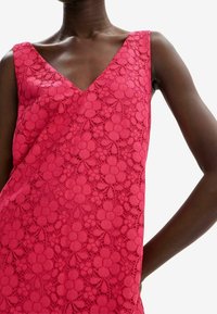 Detailní záběr modelky v růžových krajkových šatech s výstřihem do V, které zdobí květinové vzory a složité detaily na látce.