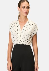 Short-sleeve blouse in cream with black polka dots, featuring a deep V-neck and gathered shoulders. Paired with high-waisted black trousers.