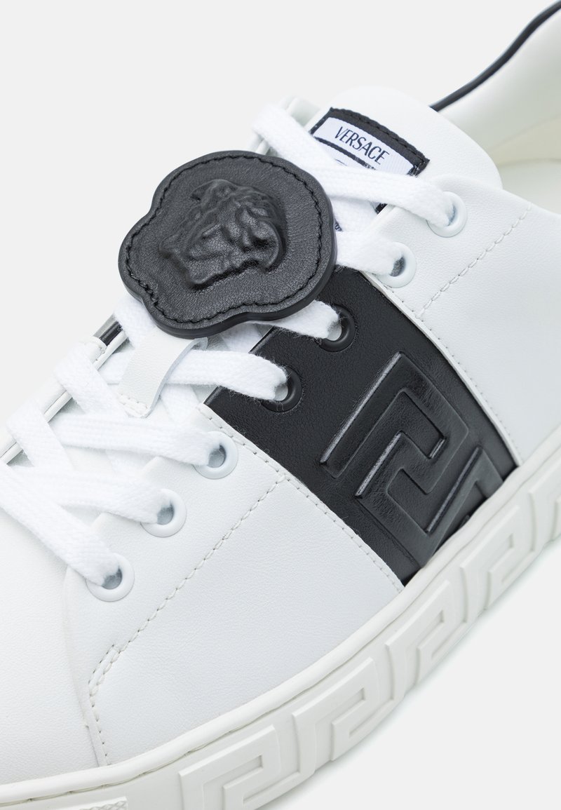 Weiße Sneaker mit schwarzen Lederakzenten, die ein erhabenes geometrisches Muster und ein schwarzes Löwenemblem im Schnürbereich aufweisen. Gummisohle.