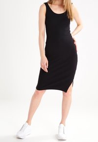 Czarna, dopasowana, bezrękawnikowa sukienka midi z okrągłym dekoltem i rozcięciem po boku; wykonana z gładkiego, elastycznego materiału. Noszona z białymi sneakerami.