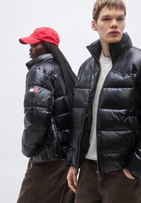 Čierne prešívané bundy s lesklým povrchom, vybavené vysokým golierom a zapínaním na zips, nosené so sivým oblečením a červenou čiapkou.