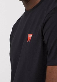 Černé bavlněné tričko s kulatým výstřihem. Na levé straně hrudi se nachází malé červené logo "Wrangler". Krátké rukávy a hladký povrch.